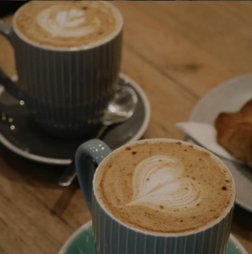 Dos capuchinos con arte latte en forma de corazón junto a un croissant