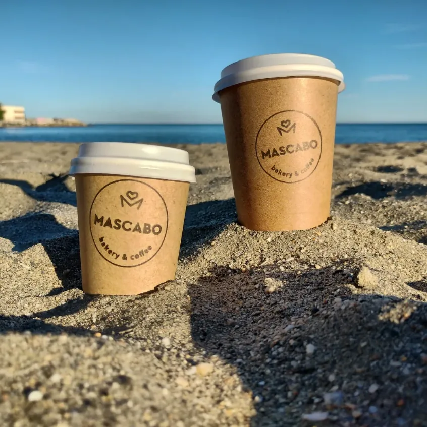 Dos vasos de café para llevar de Mascabo sobre la arena, con el mar de fondo